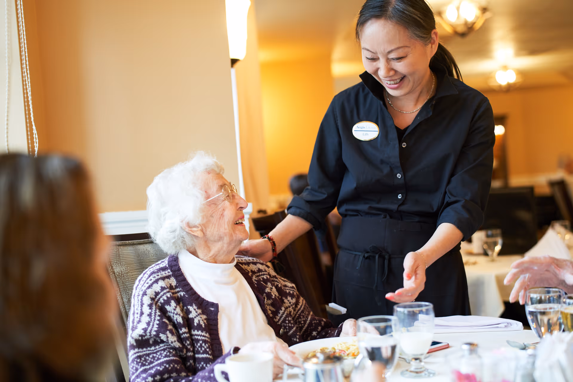 An elderly woman with white hair and glasses sitting at a dining table, smiling and looking up at a smiling female staff member wearing a black uniform and name tag. The setting appears to be a warm, well-lit dining room with tables set for a meal.