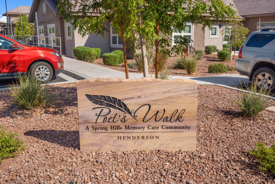 Stone sign reading "Poet's Walk A Spring Hills Memory Care Community HENDERSON" set in landscaped gravel with the facility building and parked cars behind it.