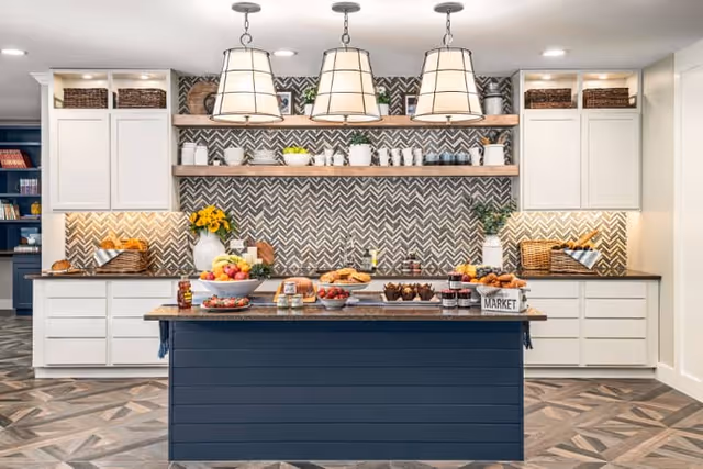 Bright communal kitchen with a central island, three pendant lights, open shelving, patterned backsplash, and food displayed on the counters.