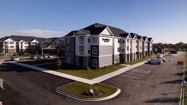 Exterior view of a multi-story senior living facility building with a parking lot and landscaped areas under a clear blue sky. The building has a combination of white and gray siding with multiple windows and a peaked roof. A few vehicles are parked in the lot, and a person is visible near the far end of the building.