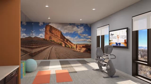 A bright exercise room with two orange yoga mats on the floor, a large exercise ball, and a stationary exercise bike facing a wall-mounted TV. The back wall features a large mural of a scenic outdoor amphitheater with red rock formations under a blue sky with clouds. The room has large windows letting in natural light and light-colored flooring.