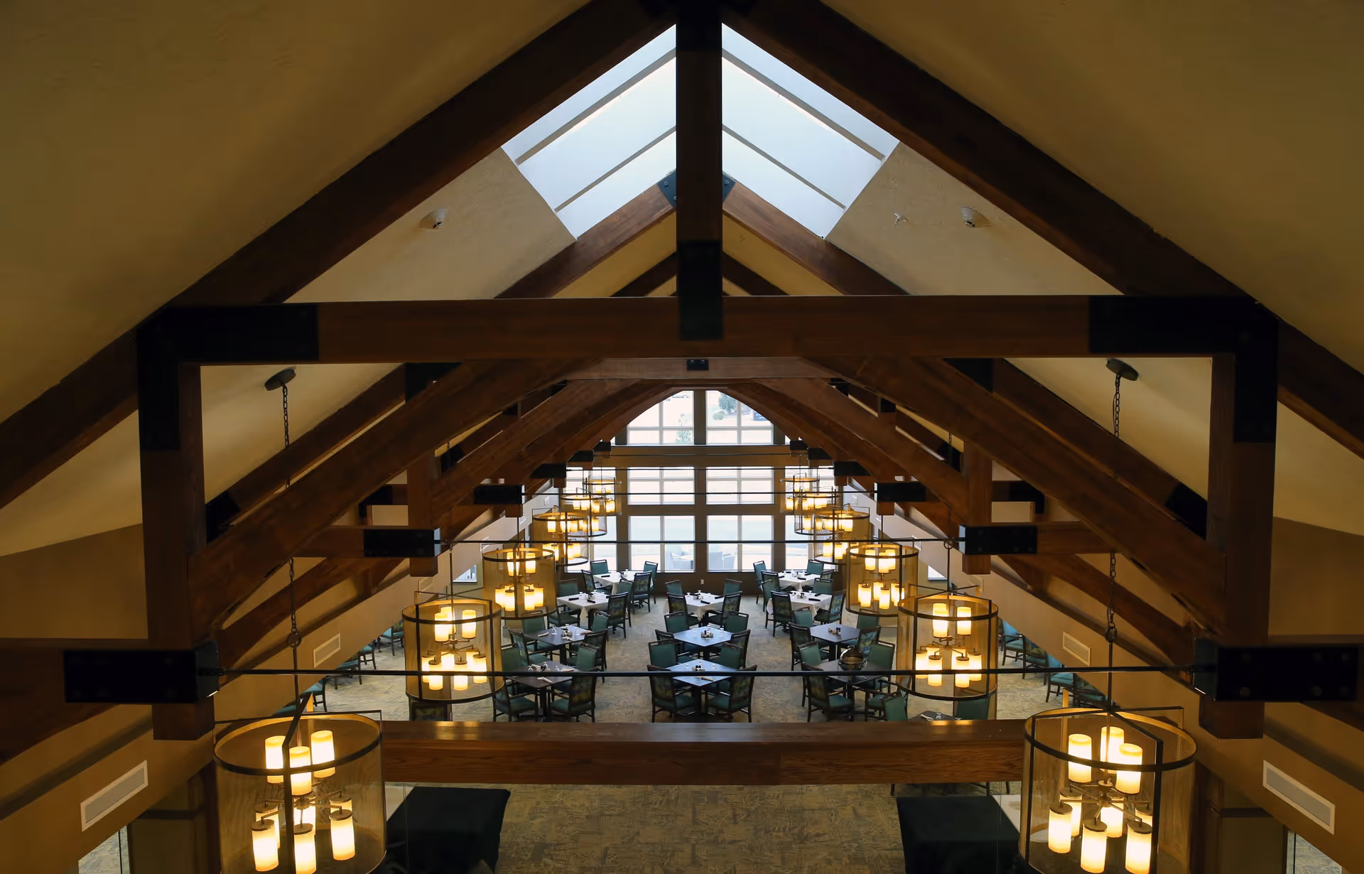 Interior view of a spacious dining room with high vaulted ceilings featuring exposed wooden beams and multiple hanging light fixtures. The room contains several tables with chairs arranged neatly, and large windows at the far end allow natural light to enter.