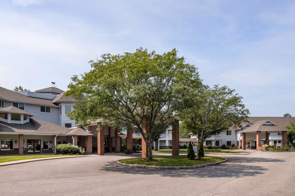 Exterior view of Anchor Bay at Pocasset senior living facility showing a driveway with a circular island featuring two large trees, surrounded by multi-story buildings with brick columns and beige siding under a partly cloudy sky.