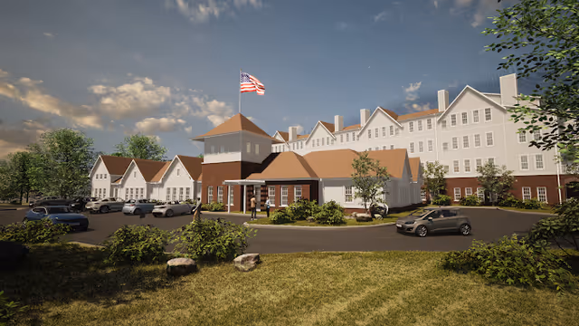 Front exterior view of The Westerly Pewaukee senior living building with an American flag, entrance drive, parked cars, and landscaped grounds.