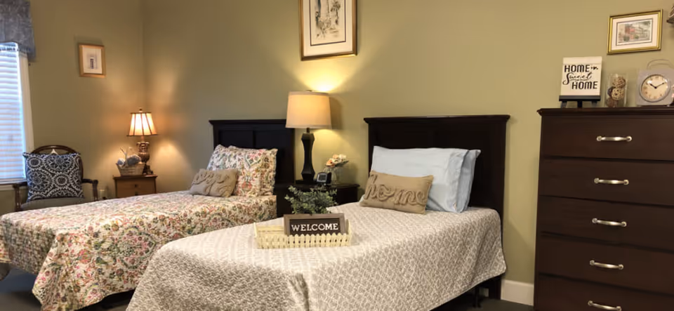 A cozy bedroom with two single beds, each with patterned bedspreads and decorative pillows. Between the beds is a nightstand with a lamp and a small plant. A wooden dresser with a clock and a 'Home Sweet Home' sign is on the right side of the room. The walls are painted light green and decorated with framed artwork.