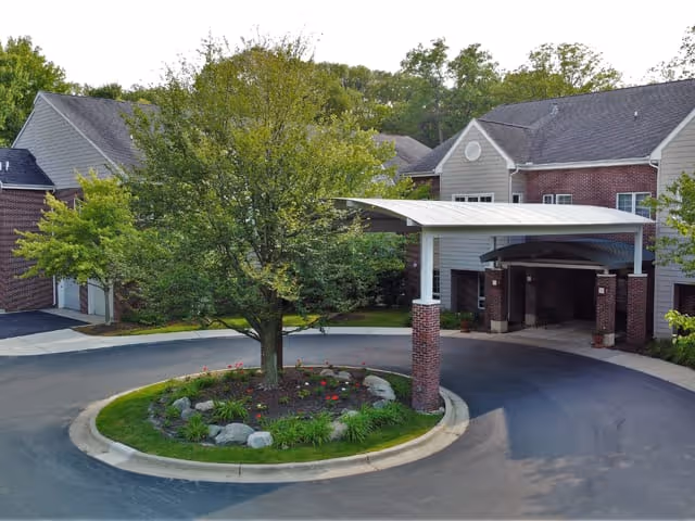 Covered porte-cochère and circular landscaped island with a tree at the front entrance of a two-story brick-and-siding building.