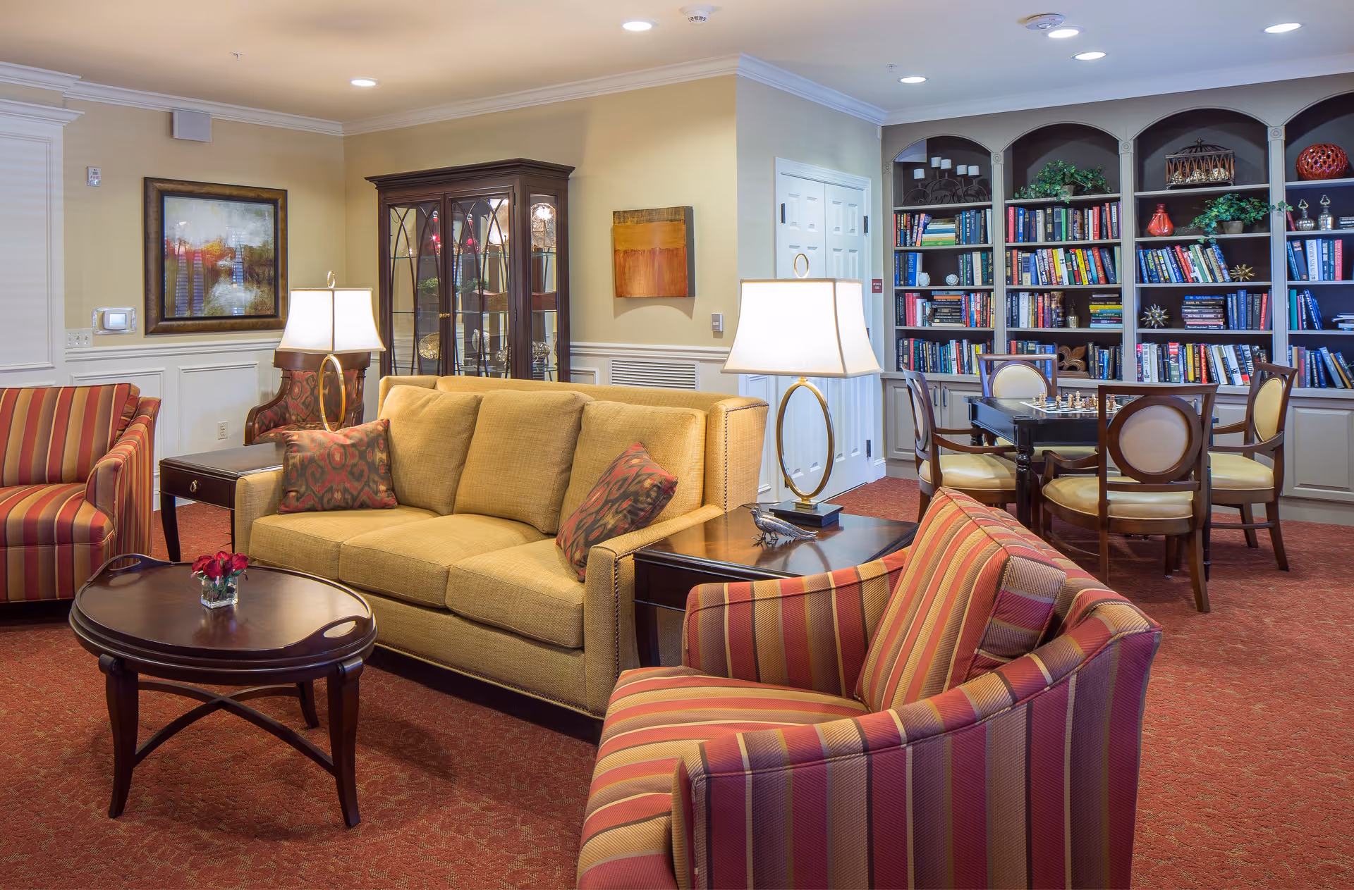 A cozy senior living common area with a yellow sofa, two striped armchairs, a wooden coffee table with a small flower vase, two table lamps, a dark wood china cabinet, and a bookshelf filled with books and decorative items. There is also a table with chairs set up for playing chess.