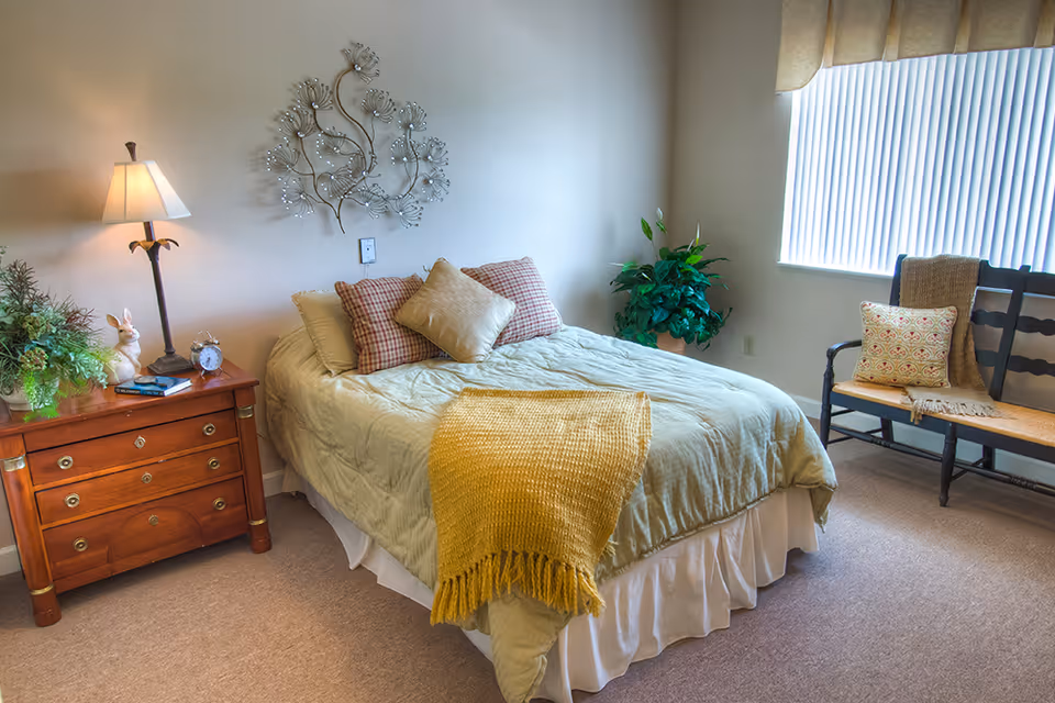 A cozy bedroom with a neatly made bed featuring a green comforter, three pillows, and a yellow throw blanket. To the left of the bed is a wooden nightstand with a lamp, a small clock, a decorative rabbit, and some greenery. On the wall above the bed is a metal floral wall decoration. To the right, there is a window with vertical blinds and a wooden bench with a cushion and a throw blanket. A potted plant is placed near the window.