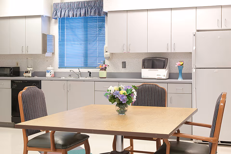 A clean and bright kitchen area with white cabinets, a refrigerator, dishwasher, microwave, blender, and a countertop with a paper towel dispenser and small appliances. In the foreground, there is a wooden table with a vase of colorful flowers and four cushioned chairs around it. A window with blue blinds and a blue valance is above the sink.
