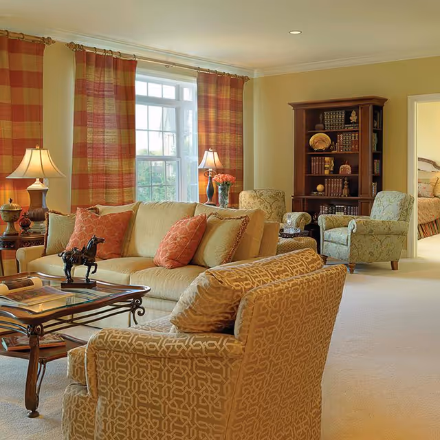 Bright living room with beige sofa and patterned armchairs around a glass coffee table, a bookshelf, and a large window with plaid curtains.