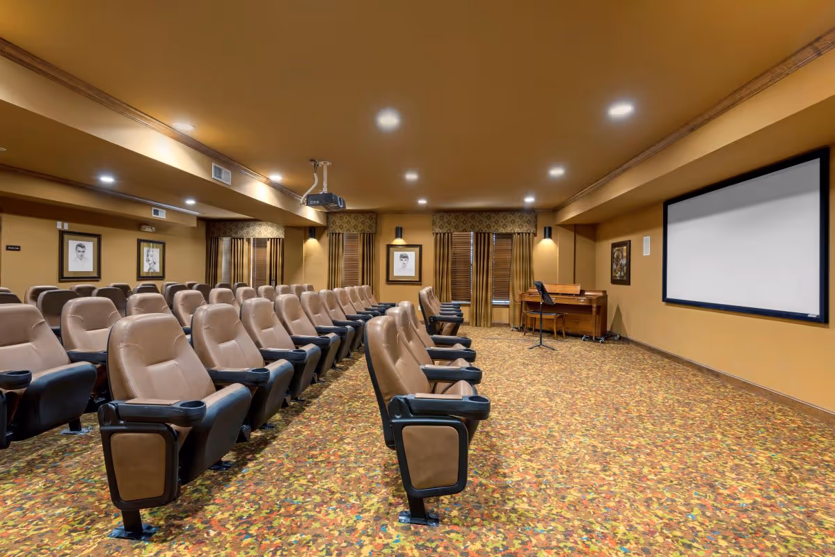 A senior living facility theater room with multiple rows of comfortable brown reclining chairs facing a large projection screen. The room has warm yellow walls, patterned carpet, framed artwork on the walls, and a piano with a music stand in the corner.