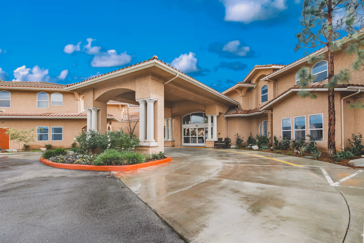 Exterior view of Ivy Park at Simi Valley senior living facility showing the main entrance with a covered driveway, beige stucco walls, red-tiled roof, landscaped flower beds, and a clear blue sky with some clouds.