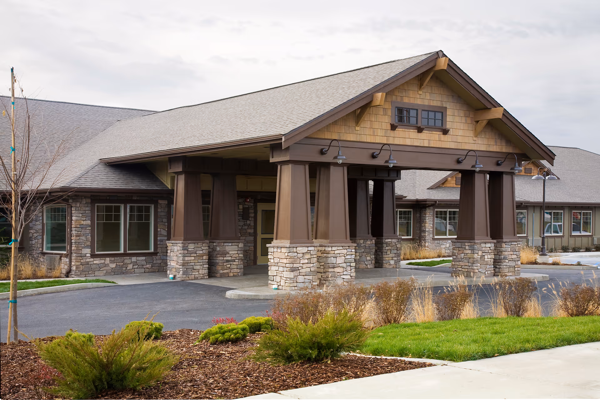 Exterior view of Creekside Inn Memory Care Community building entrance with stone pillars, wooden beams, and a covered driveway. The building has a pitched roof and multiple windows, surrounded by landscaped greenery and shrubs.