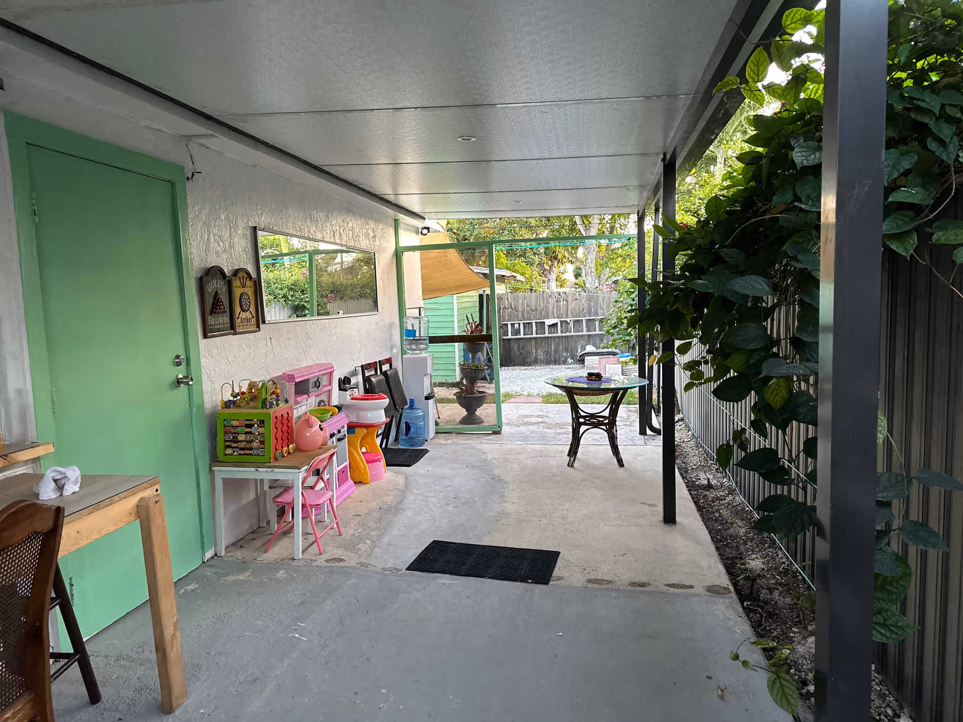 Covered outdoor patio area with a green door on the left, children's play kitchen and toys along the wall, a small table with chairs, and plants growing along the right side fence. The patio opens to a garden area with trees and a wooden fence in the background.