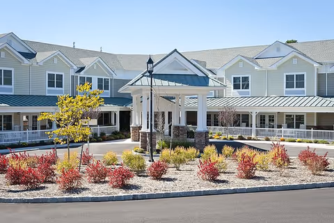 Front entrance of a two-story senior living building with a covered porte-cochere and landscaped roundabout.