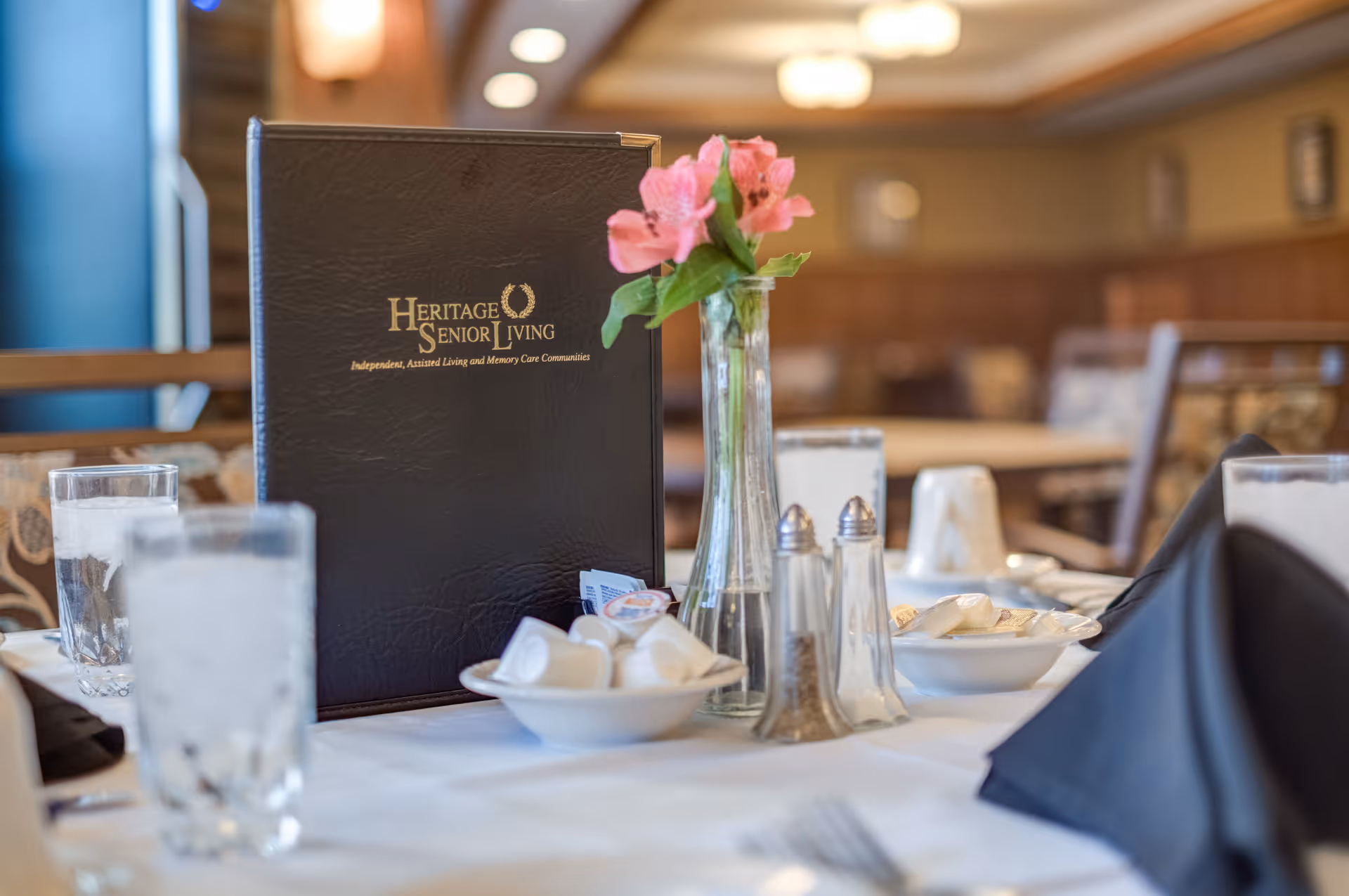 A dining table set with glasses of water, salt and pepper shakers, small bowls with creamers, a vase with pink flowers, and a black menu folder labeled 'Heritage Senior Living' in a softly lit dining room.