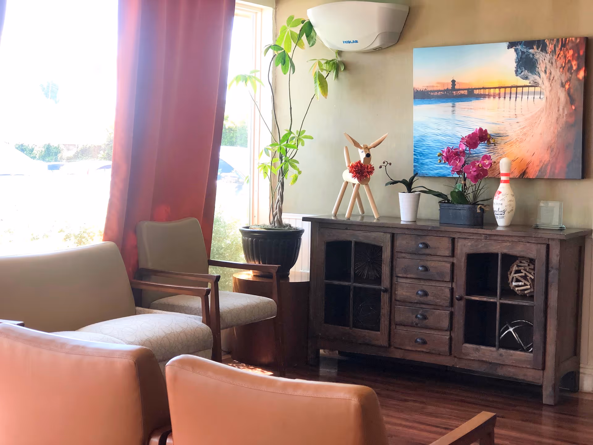 A cozy seating area with beige and light brown chairs near a large window with red curtains. A wooden cabinet against the wall holds decorative items including a potted plant, a wooden deer figurine, a bowling pin, and a vibrant pink orchid. Above the cabinet hangs a colorful photo of a wave and pier at sunset. The floor is wooden and the room is softly lit by natural light.
