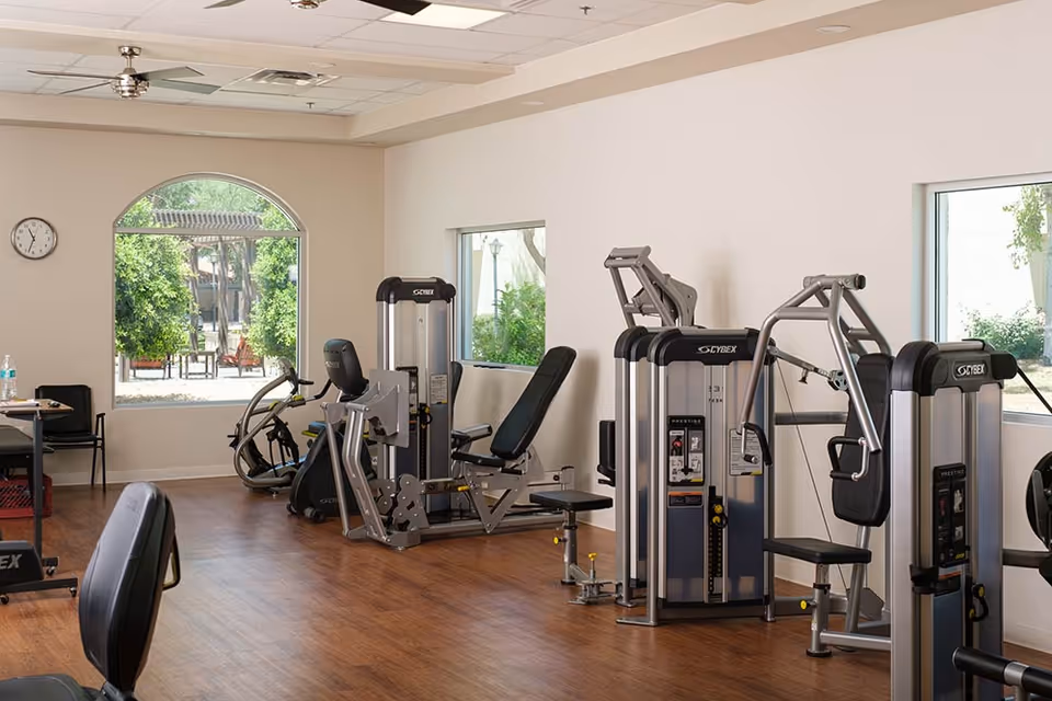 A fitness room with exercise machines including Cybex strength training equipment, a stationary bike, and a recumbent bike. The room has wooden flooring, white walls, large windows showing greenery outside, and a clock on the wall.