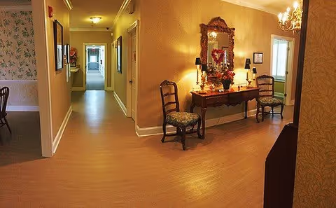Interior view of a hallway in a senior living facility with wooden flooring, two wooden chairs with cushions, a decorative table with a mirror above it, lamps, and a chandelier. The hallway leads to other rooms with visible doorways and framed pictures on the walls.