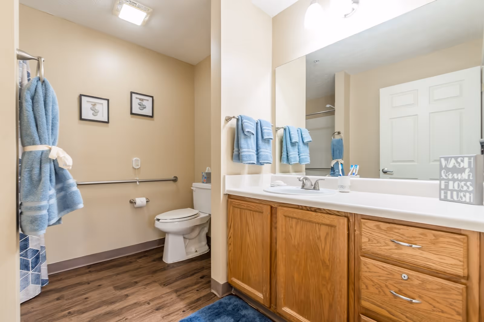 A clean bathroom with a white toilet, wooden vanity with a white countertop and sink, a large mirror above the sink, blue towels hanging on the wall and a towel rack, two framed pictures on the beige wall, and a blue bath mat on the wooden floor.