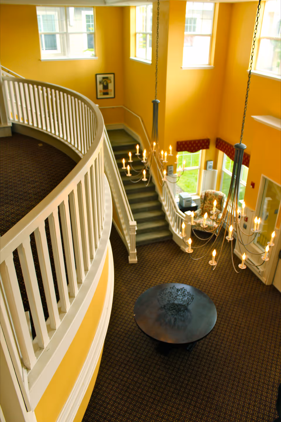 Interior view of a senior living facility with yellow walls, a curved white railing on the upper floor, a staircase leading down to a carpeted area with a round black table, two chandeliers with lit candles, large windows letting in natural light, and a floral armchair near the entrance.