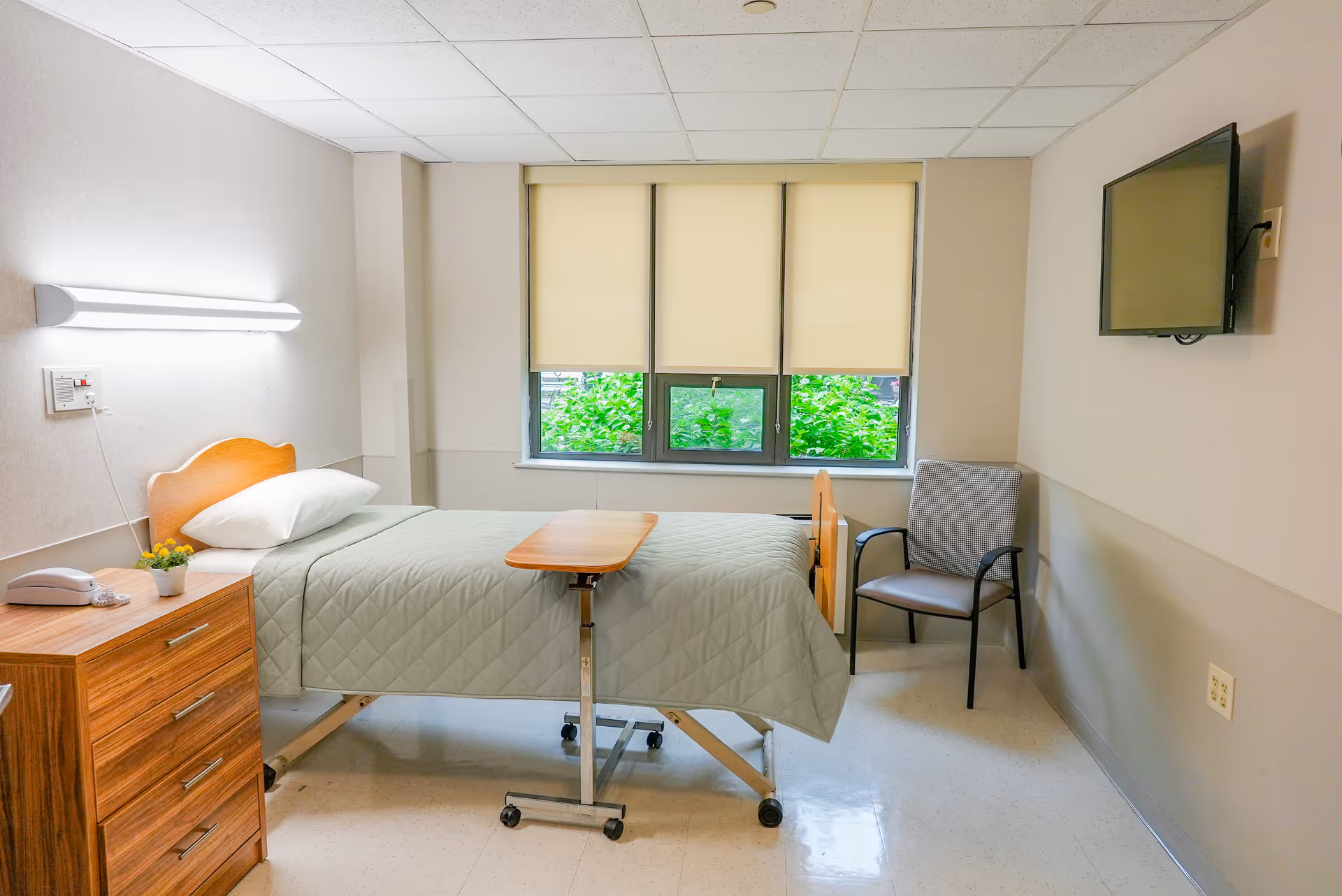 A tidy nursing facility bedroom with a single hospital bed, overbed table, wooden dresser, chair, wall-mounted TV and a window with shades.