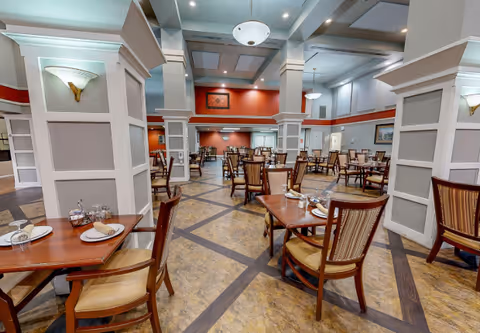 Spacious dining room with multiple wooden tables and chairs, high coffered ceilings, columns, and place settings.