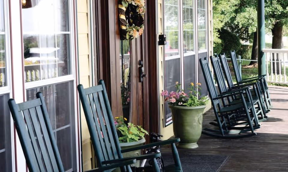 A wooden porch with several dark green rocking chairs lined up along the side of a yellow building. There are large windows and a wooden door decorated with a floral wreath. Two green flower pots with pink and yellow flowers are placed near the door. Trees and greenery are visible in the background.