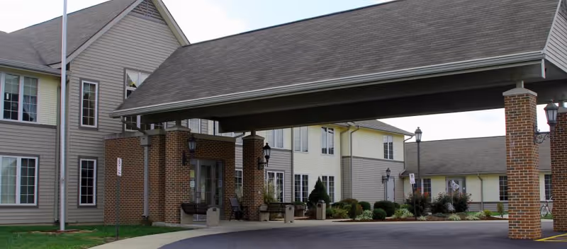 Covered porte-cochère and main entrance of a senior healthcare building with brick columns, windows, and a landscaped driveway.