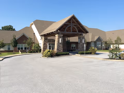 Front entrance of a single-story senior care building with a covered porte-cochere and surrounding parking area under a clear blue sky.