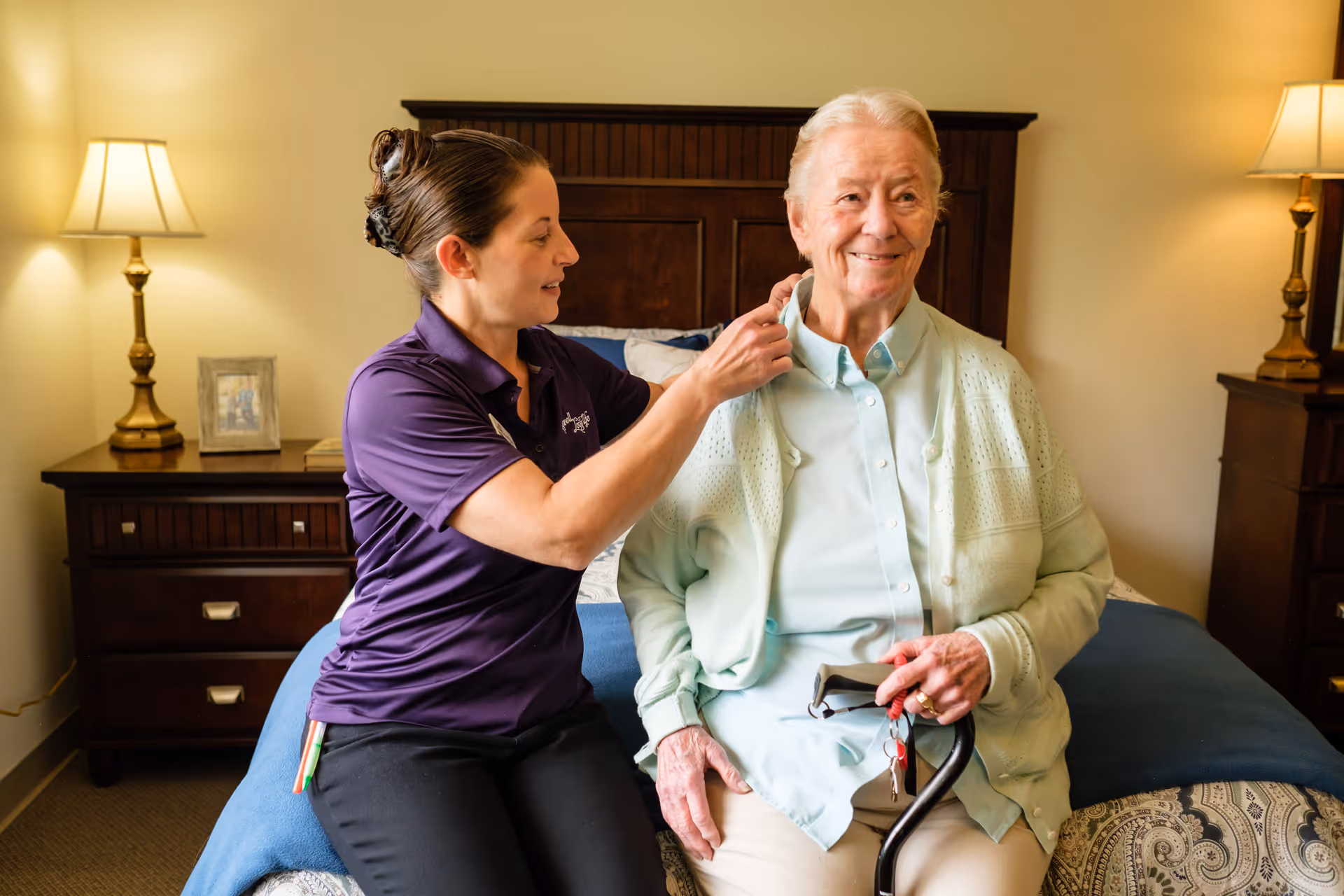 A caregiver in a purple shirt is helping an elderly woman button her light green cardigan while sitting on a bed in a warmly lit bedroom with wooden furniture and lamps on either side.