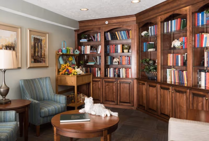 Cozy common room with wooden built-in bookshelves, upholstered chairs, and a round coffee table with a small white stuffed animal.