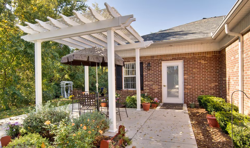 Outdoor patio area at Chestnut Glen Senior Living featuring a white pergola with a table and chairs underneath, surrounded by potted plants and garden beds with flowers. The patio is adjacent to a brick building with a white door and window.