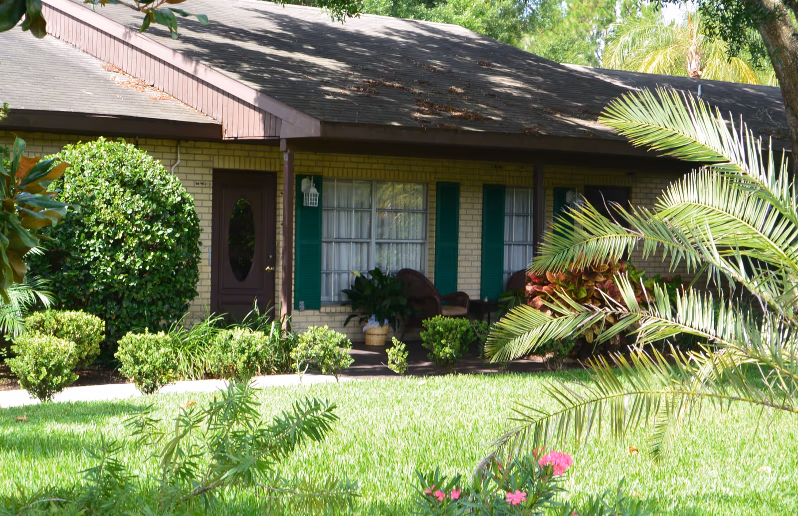 Exterior view of a single-story brick building with a brown roof, green shutters, and a brown door. The front porch has two chairs and some potted plants. The foreground features a well-maintained lawn with various bushes, plants, and a palm tree.