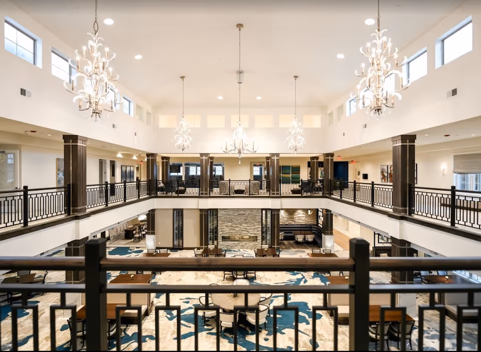 Spacious two-story common area in a senior living facility with multiple chandeliers hanging from a high ceiling. The lower level has tables and chairs arranged on a patterned carpet, and the upper level features a balcony with railings overlooking the area below. The walls are light-colored with large windows near the ceiling allowing natural light to enter.