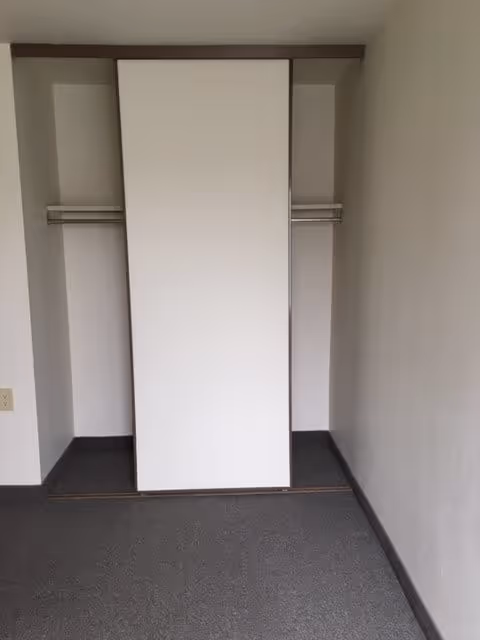 Empty closet with a sliding door partially open, showing a hanging rod inside. The closet is in a room with plain white walls and gray carpeted floor.