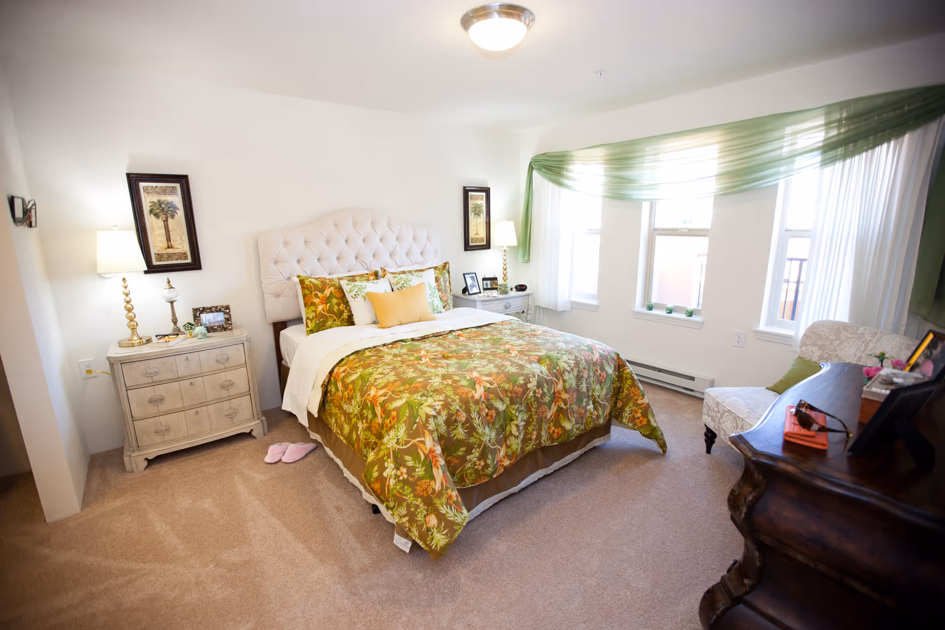 A bright and cozy bedroom with a large bed featuring a tufted white headboard and floral green bedding. There are two matching nightstands with lamps and framed pictures on either side of the bed. The room has beige carpet, three windows with sheer white curtains and a green swag valance, a patterned armchair, and a dark wooden dresser with personal items on top.