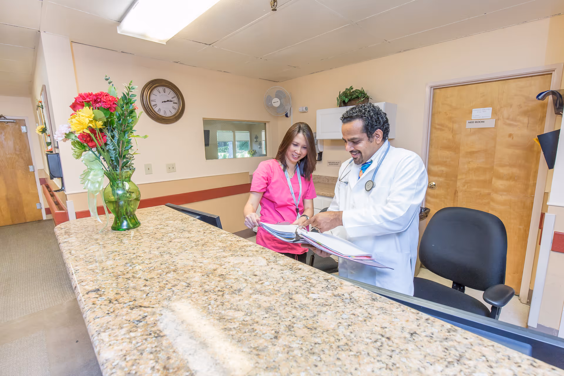 A medical professional in a white coat and a staff member in pink scrubs are standing behind a granite reception desk, looking at documents together. There is a vase with colorful flowers on the desk, a wall clock, and a door labeled 'MED ROOM' in the background.