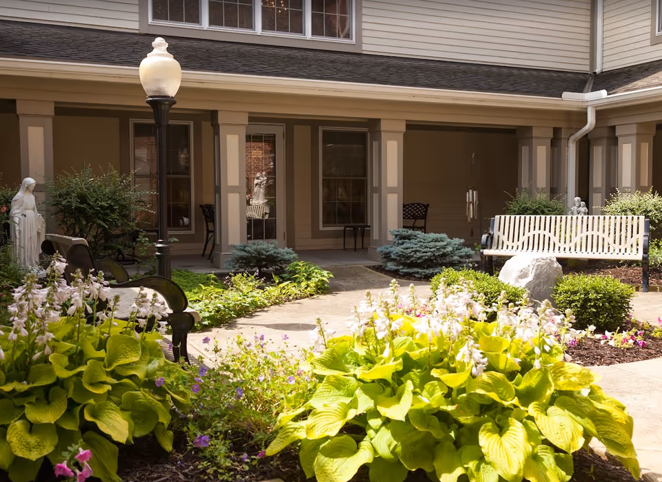 Sunny courtyard garden with benches, flowering plants, statues, and a covered walkway in front of the building.
