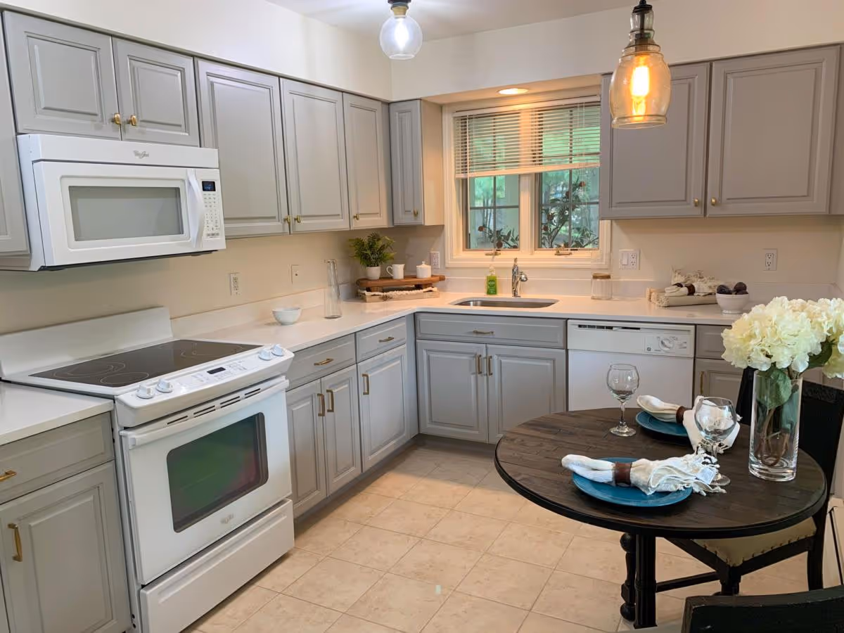 A modern kitchen with light gray cabinets, a white electric stove with oven, a white microwave above it, and a dishwasher. There is a window above the sink with blinds partially open, and a small round dining table set for two with blue plates, napkins, wine glasses, and a vase of white flowers. The kitchen has tiled flooring and two hanging pendant lights.