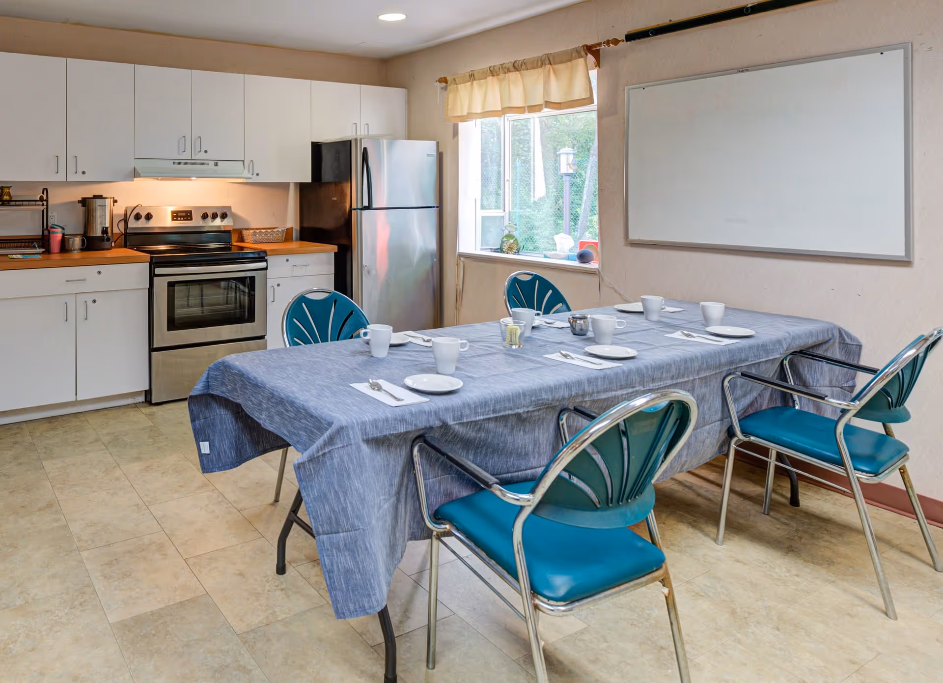 A kitchen and dining area with a table covered by a blue tablecloth set with white plates, cups, and utensils. The kitchen features white cabinets, a stainless steel oven, and a refrigerator. A window with a beige valance lets in natural light, and a whiteboard is mounted on the wall.