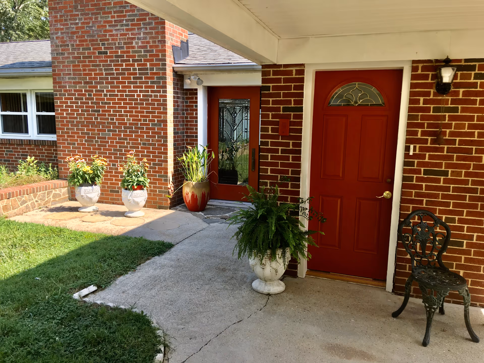 Covered brick entryway with a red door, decorative glass door, potted plants, and a metal chair on a concrete walkway.