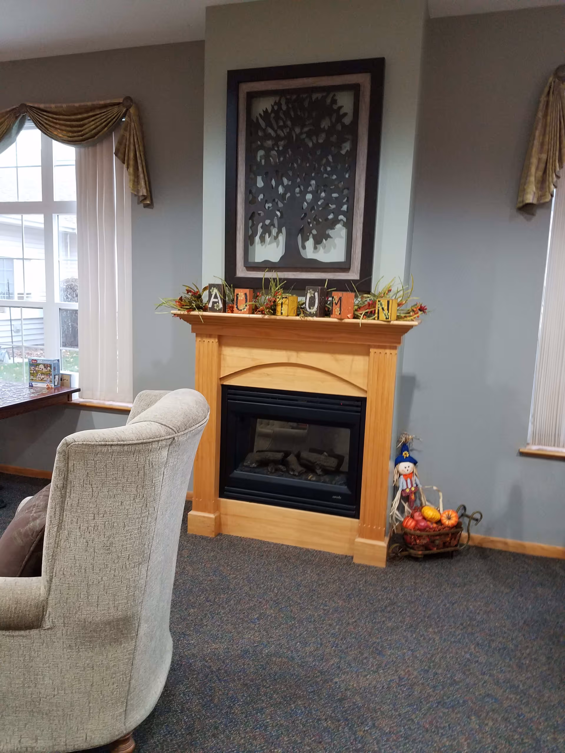 A cozy living room area with a light gray upholstered armchair facing a wooden fireplace mantel decorated with autumn-themed blocks and foliage. Above the mantel hangs a framed metal tree artwork. To the right of the fireplace is a small decorative scarecrow figure with a basket of pumpkins and gourds. The room has gray walls, carpeted floor, and windows with vertical blinds and gold valances.