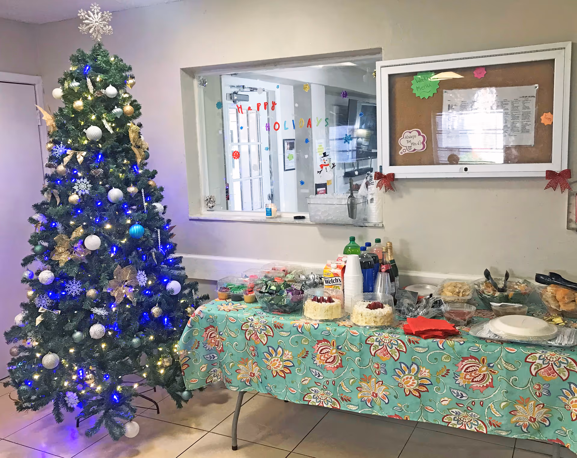 A decorated Christmas tree with blue and white lights and ornaments stands next to a table covered with a floral tablecloth. The table holds various food items, drinks, disposable cups, plates, and napkins. Behind the table is a window with holiday decorations and a bulletin board with papers and festive bows on the wall.