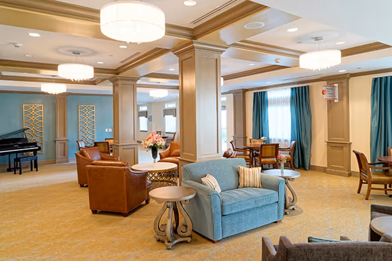 A spacious and elegant common area in a senior living facility featuring comfortable seating including a blue loveseat with striped pillows, brown leather armchairs, round wooden side tables, and dining tables with chairs near windows with teal curtains. The room has beige carpet, coffered ceilings with recessed lighting and chandeliers, decorative columns, and a black grand piano in the background.