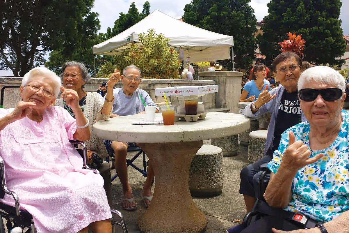 A group of elderly people sitting around a round outdoor concrete table with drinks and pizza boxes, enjoying a sunny day at an outdoor gathering with trees and a white canopy tent in the background.