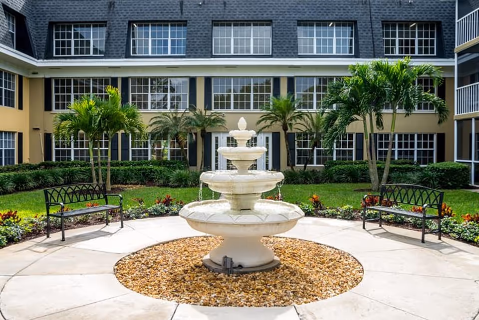 Courtyard of a senior living facility featuring a three-tier stone fountain, two benches, palm trees, and surrounding building windows.