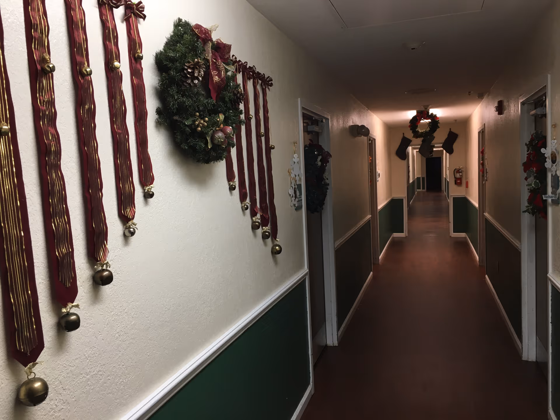 A long indoor hallway decorated with Christmas wreaths, red ribbons with gold bells, and hanging stockings. The walls are painted white on the upper half and green on the lower half, with a wooden floor and several doors along the corridor.