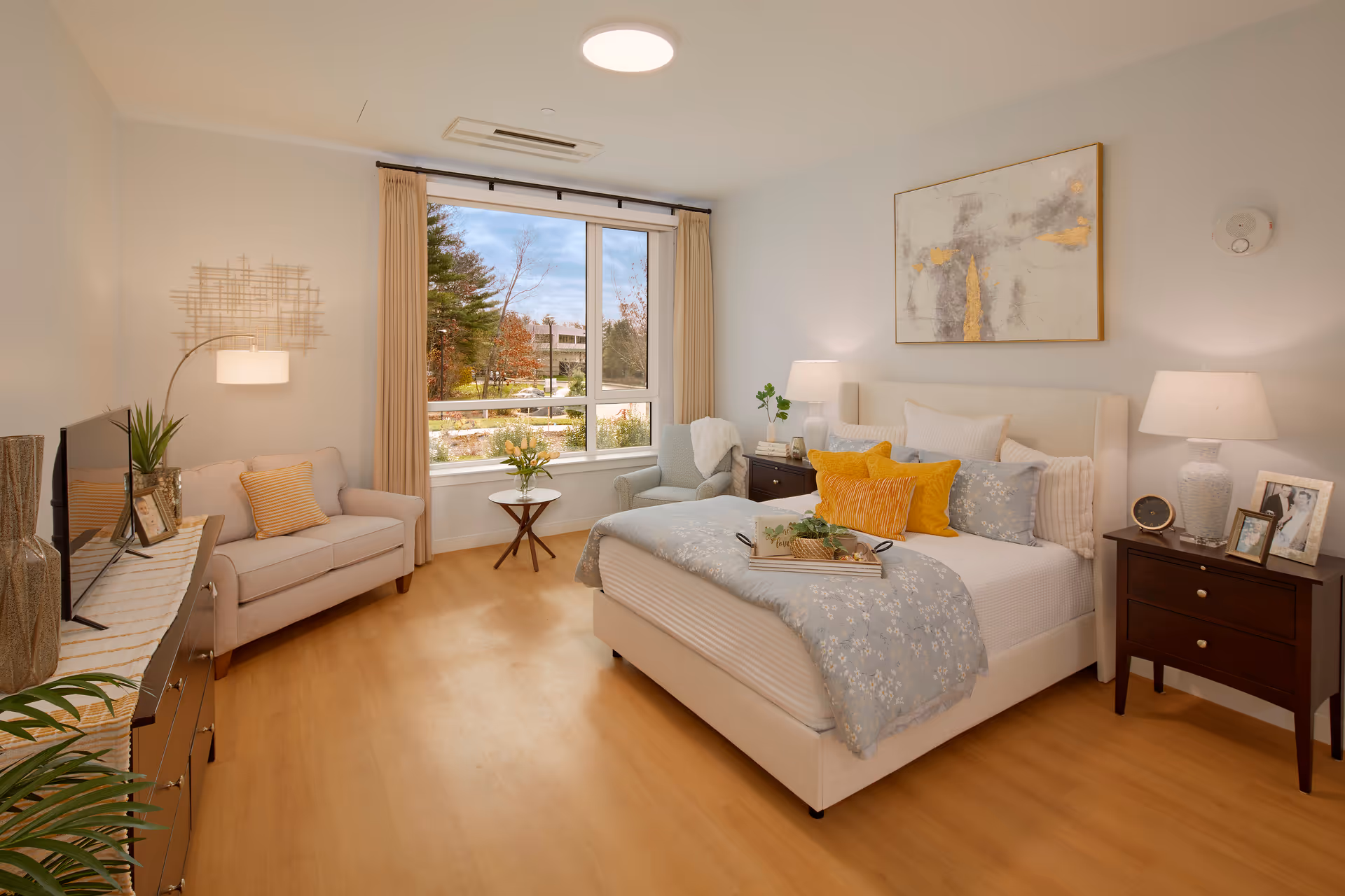 A bright and cozy bedroom with a large window showing an outdoor view. The room features a comfortable bed with multiple pillows and a floral bedspread, two dark wooden nightstands with lamps and framed photos, a light blue armchair with a white throw, a small round side table with a vase of flowers, a beige loveseat with a striped pillow, a dresser with a TV and decorative items, and light wood flooring.