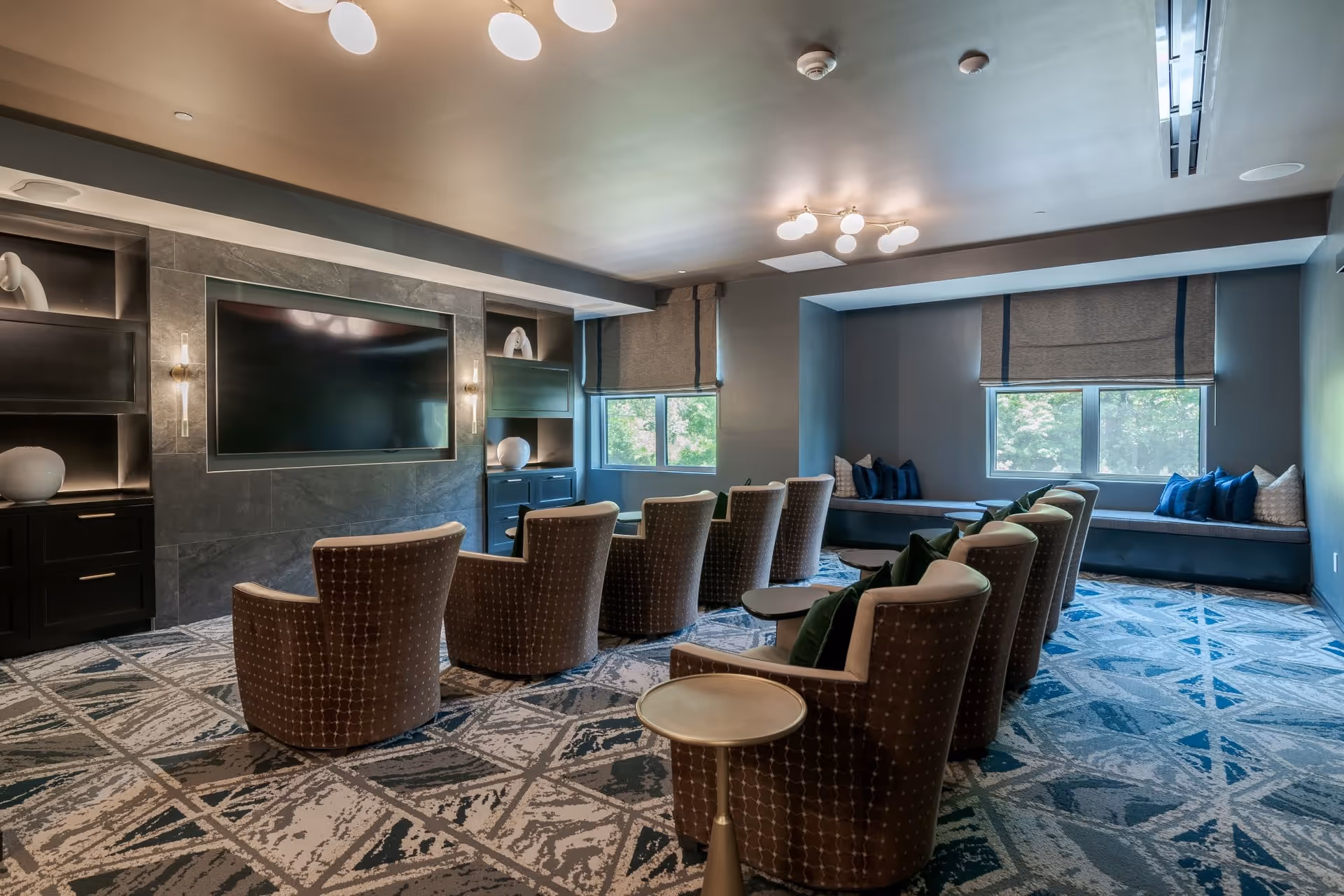 A cozy media room with upholstered theater-style chairs facing a large wall-mounted TV, side tables, and window seating.